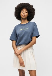 Γυναίκα με σγουρά μαλλιά που φοράει μπλε μπλουζάκι Nike Athletics Club και λευκή πλισέ φούστα, στέκεται με σταυρωμένα χέρια.