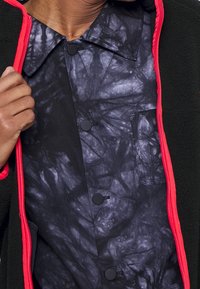 Fleece bunda s černým interiérem, která má strukturovaný, abstraktní vzor, černé knoflíky a jasně červený zip a lemování.