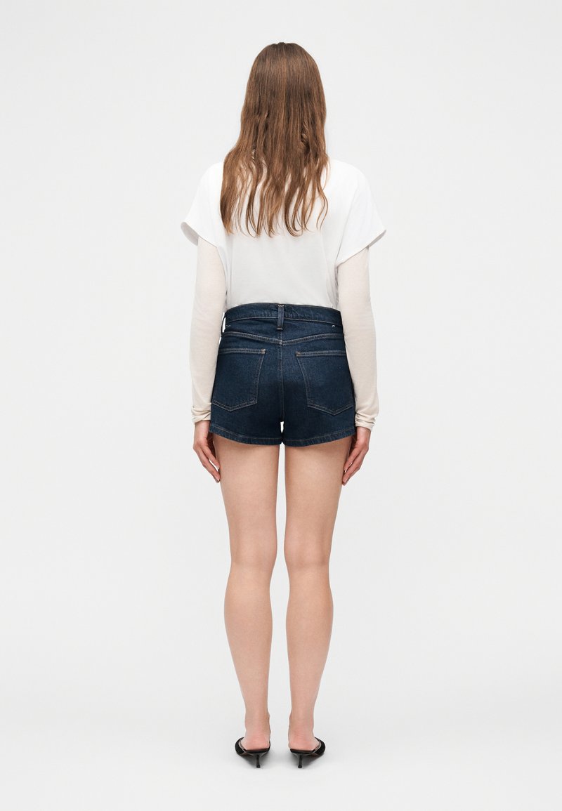 Femme aux longs cheveux portant des hauts blancs superposés, un short en denim foncé et des mules à talons noires, debout de dos sur un fond uni.
