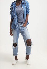 Jeansjacka med huva, blå T-shirt, ljusblå slitna jeans och vita slip-on skor. Har slitet tyg och uppvikta ärmar.