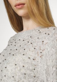 Frau mit langen blonden Haaren, die einen hellgrauen Strickpullover trägt, der über der Brust und den Schultern mit kleinen metallischen Perlen verziert ist.