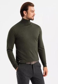 Pull à col roulé vert olive au design ajusté, fabriqué en matériau doux, avec des manches longues et des poignets côtelés. Associé à un pantalon gris foncé.