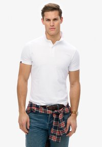 Vit kortärmad polo med krage, matchad med blå jeans och en röd rutiga flanellskjorta knuten runt midjan. Brunt bälte och klocka.