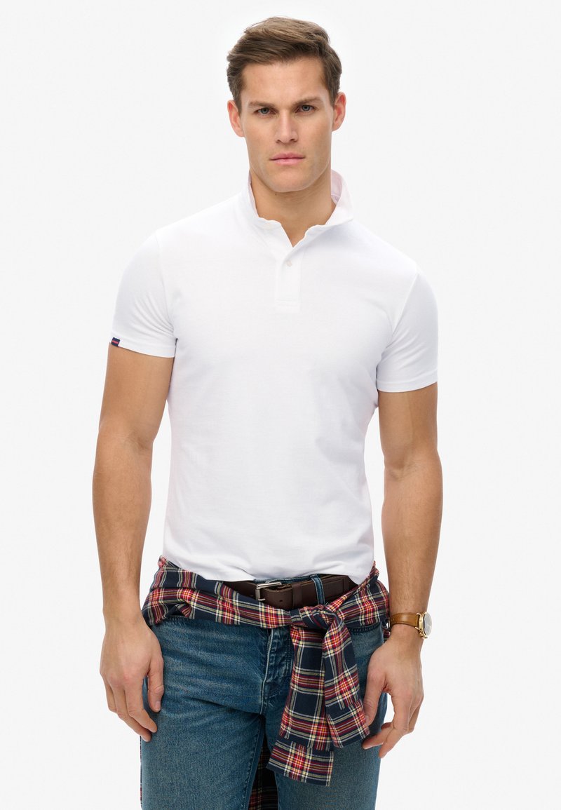 Vit kortärmad polo med krage, matchad med blå jeans och en röd rutiga flanellskjorta knuten runt midjan. Brunt bälte och klocka.