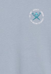 Una tela de color azul claro presenta un logotipo circular bordado en blanco con un diseño de paraguas cruzados y un sombrero, etiquetado "Hackett London".