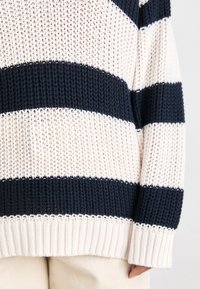 Pull en maille avec larges rayures horizontales bleu marine et blanches et poignets côtelés, porté avec un pantalon beige clair, bras détendu sur le côté.