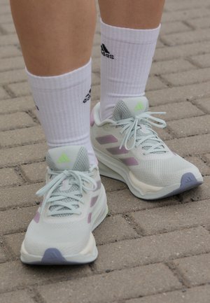 Osoba nosząca białe buty sportowe Adidas z pastelowo fioletowymi akcentami oraz białe skarpetki Adidas stoi na szarej brukowanej nawierzchni.
