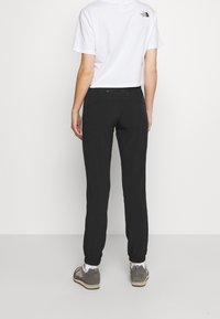 Joggers noirs à coupe décontractée avec poignets élastiques, associés à un t-shirt blanc court avec un petit logo au dos. Portés avec des chaussures grises.