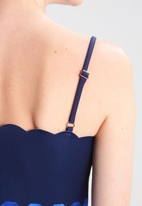 Námořnická modrá blůza se vykrojeným výstřihem, nastavitelné ramínka a výrazný vrstvený lem v různých odstínech modré. Hladká textura.