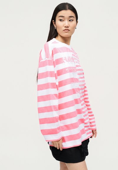 Νεαρή γυναίκα με μακριά σκούρα μαλλιά, που φοράει μακρυμάνικη ροζ και λευκή ριγέ μπλούζα και μαύρη φούστα, κοιτάζει πλάγια.