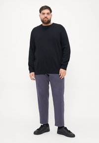 Pulover negru tricotat cu guler rotund, asortat cu pantaloni purpura largi și adidași negri, stând pe un fundal alb.
