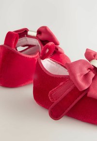 Rote Samt-Baby-Schuhe mit einer strukturierten Oberfläche, abgerundeter Zehenpartie und großen rosa Schleifenakzenten auf der Vorderseite. Sie verfügen über ein unkompliziertes Slip-On-Design.