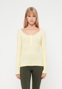 Pitkähiha, ribattu paita vaaleankeltaisena, jossa on avara kaula-aukko ja viiden napin lista. Tiukka malli täydentää vihreitä housuja.