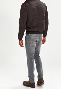 Polstret sort bomberjakke med ribbet kant, parret med lysegrå jeans og brune læderstøvler, set fra ryggen.