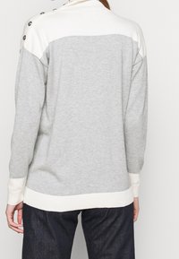 Grå tröja med krämfärgade detaljer, med hög krage, ribbade ärmslut och knapppars på axeln. Mjuk textur, avslappnad design.