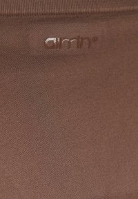 Smešasta smeđa tkanina finog tekstura, s malim reljefnim logotipom u odgovarajućoj smeđoj boji, smještenim u gornjem lijevom kutu.