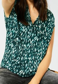 Blusa a maniche corte in teal con motivo astratto bianco, caratterizzata da un design a V e una vestibilità comoda, realizzata in tessuto morbido.