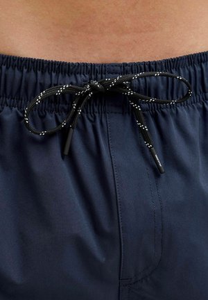 Granatowe szorty pływackie z elastycznym pasem i czarnym sznurkiem do regulacji, ozdobionym białymi plamkami. Gładki, lekki materiał z widocznymi przeszyciami.