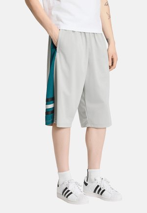 Persoon draagt grijze en blauwgroene lange shorts met zijstrepen, witte sokken en witte sneakers met zwarte strepen, staand met één hand in de zak.