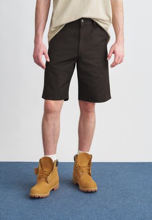 Καφέ σορτς με ίσια κοπή, κατασκευασμένα από βαμβάκι. Συνδυάζονται με σκουρόχρωμα μποτάκια αστραγάλου με κορδόνια και κίτρινες λεπτομέρειες.