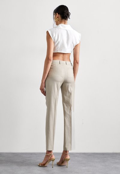 Chemisier blanc sans manches, pantalon beige ajusté et talons dorés à lanières. Texture de tissu lisse, design minimaliste, détails visibles dans le dos.