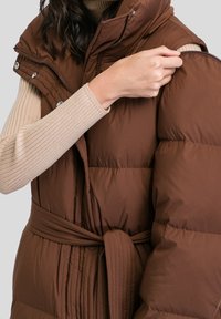 Person som bär en beige ribbad tröja och öppnar det avtagbara ärmet på en brun vadderad ärmlös jacka med bälte.