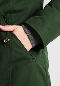 Grøn jakke med en struktureret, ribbet grå manchet. Har en sidelomme med en sølvknap som accent. Holdbart, glat stof.