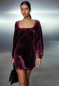 Burgundské sametové minišaty s dlouhými nařasenými rukávy, čtvercovým výstřihem a bočním rozparkem. Doplněno černou látkovou psaníčkem.