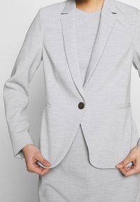 Personne portant un blazer gris clair avec un seul bouton foncé, tenant les bords du blazer près de la taille, sur un fond uni clair.