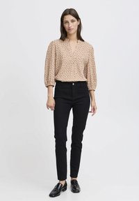Blouse beige avec des manches bouffantes et un motif abstrait noir, associée à un pantalon skinny noir et des mocassins noirs. Encolure simple et silhouette ajustée.