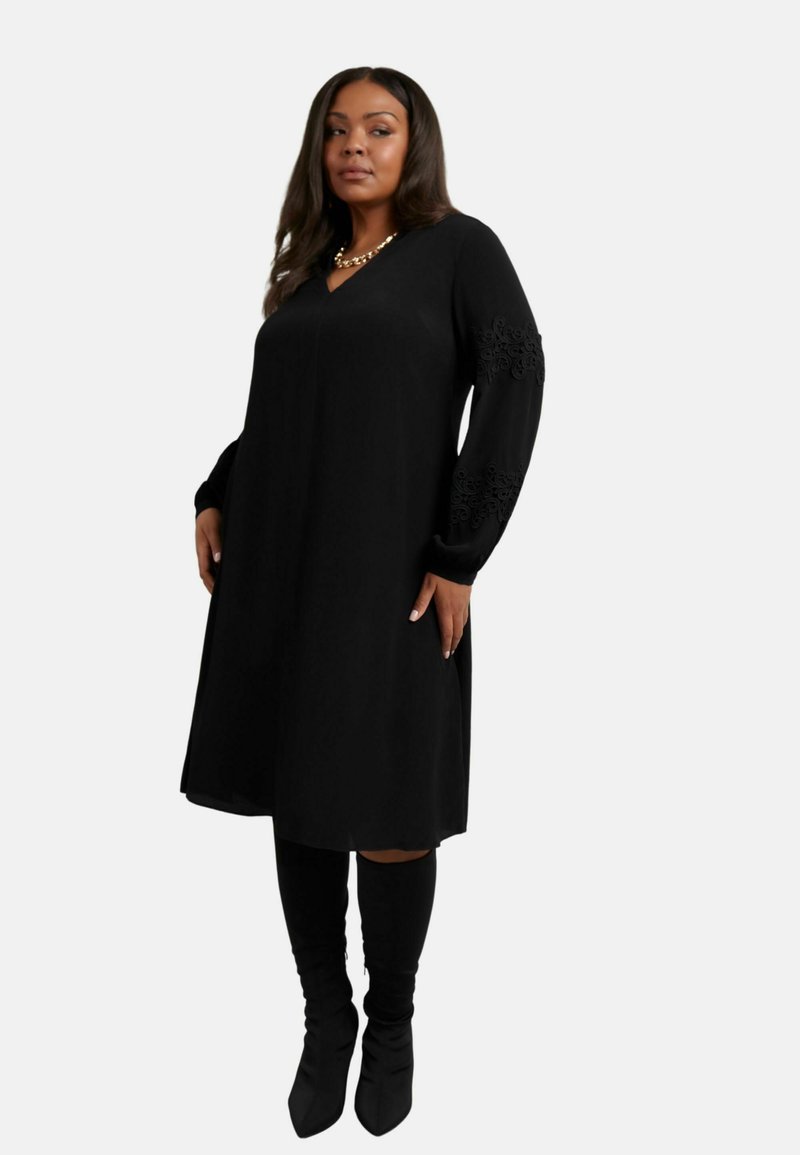 Černé šaty s výstřihem do V a dlouhými rukávy, zdobené květinovou krajkou na zápěstí. Měkká látka, po kolena, doplněné černými vysokými kozačkami.