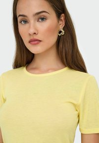 Młoda kobieta z długimi brązowymi włosami, nosząca złote rzeźbione kolczyki w kształcie obręczy oraz jasnożółtą bluzkę z krótkim rękawem, na jednolitym tle.