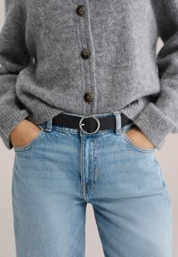 Handen in de zakken, een lichtblauwe spijkerbroek met een zwarte riem en een grijze wollen trui met knopen dragend.