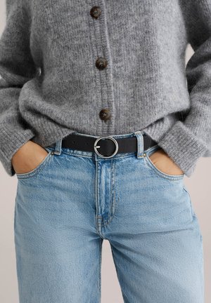 Ruky v kapse, oblečené v svetlomodré džínsy s čiernym opaskom a sivý zapínaný vlnený sveter.