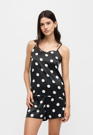Jeune femme portant un caraco en satin noir et un short à pois blancs, debout devant un fond clair uni.
