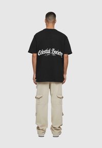 Sort bomulds T-shirt med teksten "Celestial Society" i hvid på ryggen. Samlet med cremefarvede cargo bukser og sorte sneakers.