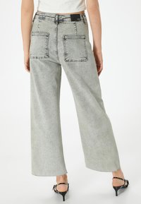 Lichtgrijze denimbroek met wijde pijpen, twee achterzakken, een franjes onderkant en een zwart merklabel op de tailleband. Soepele textuur.