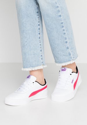 Person trägt weiße Puma-Sneaker mit pinken Streifen und lila Logo, kombiniert mit hellblauen Jeans mit ausgefranstem Saum.