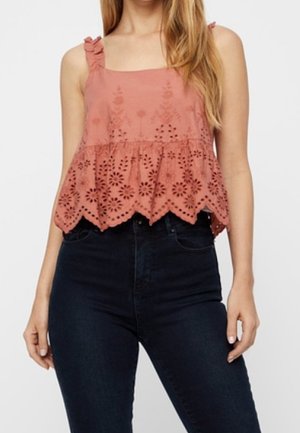 Kvinde iført en ærmeløs støvet rosa øjelysebluse med flæse stropper og mørke højtaljede jeans.