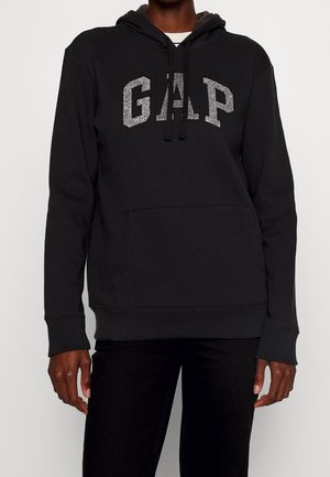 Personne portant un sweat à capuche noir avec des lettres argentées brillantes formant "GAP" sur la poitrine et un pantalon noir, debout devant un fond uni.