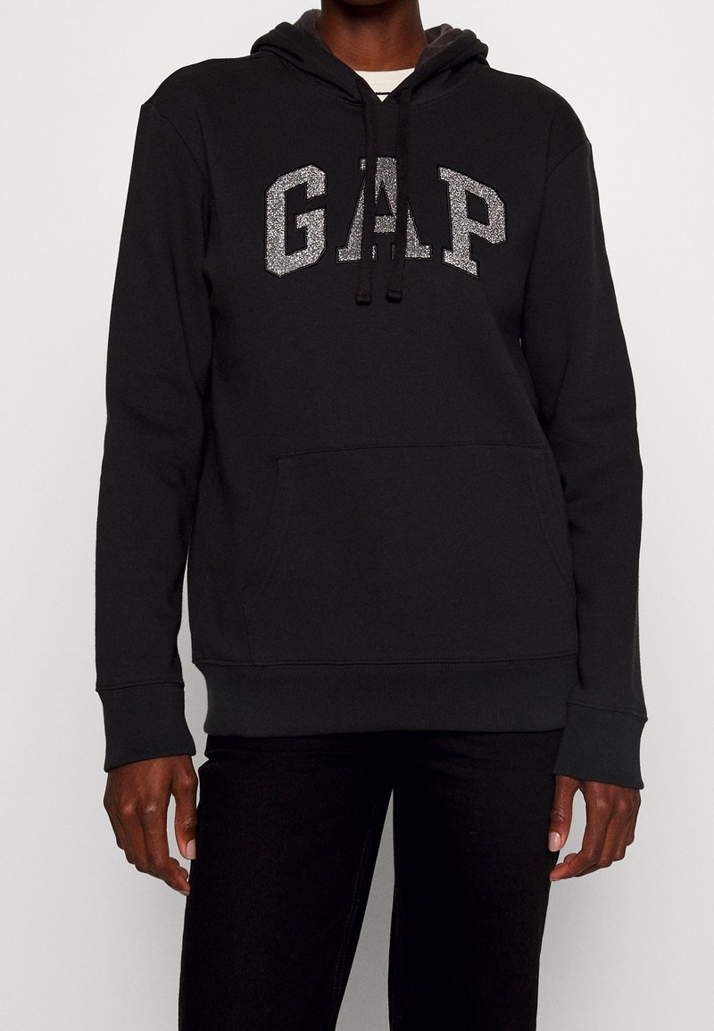 Personne portant un sweat à capuche noir avec des lettres argentées brillantes formant "GAP" sur la poitrine et un pantalon noir, debout devant un fond uni.