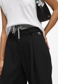 Γυναίκα με μαύρους φαρδύς παντελόνες, με ένα μικρό καρτούν αξεσουάρ, άσπρη αμάνικη μπλούζα και κρατώντας μια μαύρη τσάντα με πράσινα διακοσμητικά μαργαρίτα.