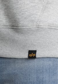Sudadera gris con puños y dobladillo acanalados. Presenta una etiqueta negra con un detalle de logo dorado. La tela parece suave y texturizada.