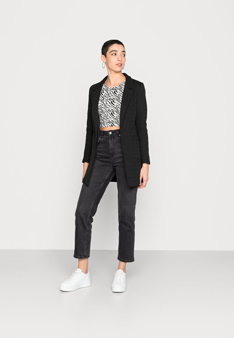 Cappotto nero su misura indossato sopra un crop top a strisce zebrate bianche e nere, abbinato a jeans neri a vita alta e sneaker bianche con lacci.