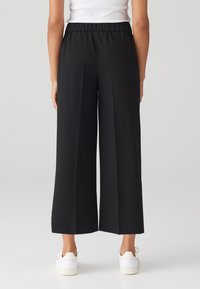 Pantalons larges noirs avec une taille élastique, texture lisse et une poche arrière unique, associés à des baskets blanches.