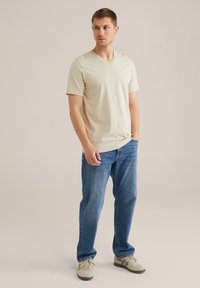 Beige V-veida T-krekliņš apvienots ar ziliem džinsiem un gaiši pelēkiem sporta apaviem. Vienkāršs dizains, īsas piedurknes un brīvs piegriezums.