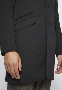 Manteau noir ajusté avec poches à rabat et fentes, boutonné devant, porté sur un pantalon à carreaux, mains le long du corps sur un fond uni.