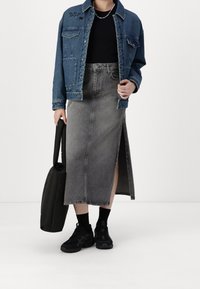 Jeansjacke mit Taschendetails, kombiniert mit einem grauen, knielangen Rock mit einem seitlichen Schlitz. Schwarze Sneakers und eine schwarze Handtasche runden das Outfit ab.