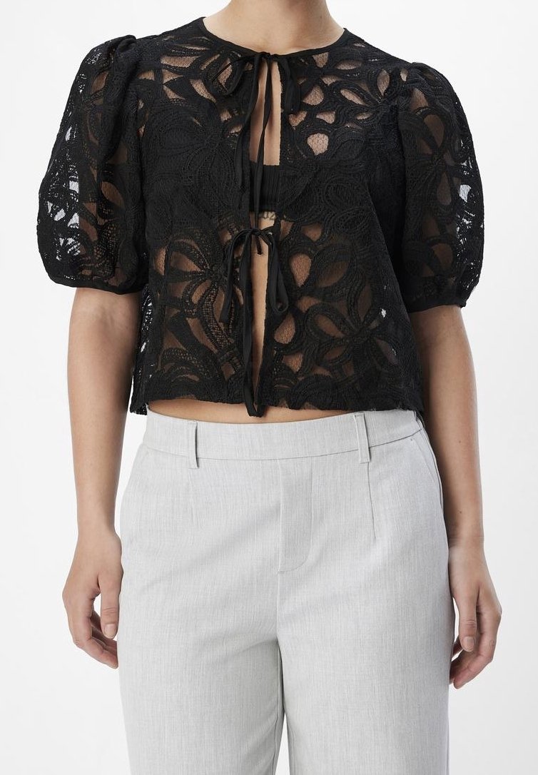 Blouse courte en dentelle noire avec manches bouffantes et fermetures à nouer, associée à un pantalon gris clair taille haute et ajusté.