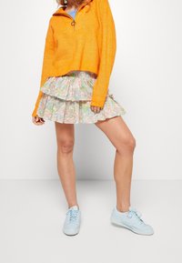 Pull en tricot orange avec un col zippé; jupe pastel à volants superposés; baskets bleu clair. Matériaux texturés et couleurs vives.